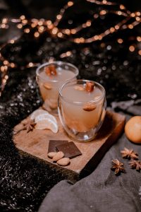 2 glasses full of an alcholic cocktail sit on a small wood block. Also on the wood block are an array of chocolate bars, almonds and orange slices. This is set before Christmas lights and garland that are slightly out of focus.